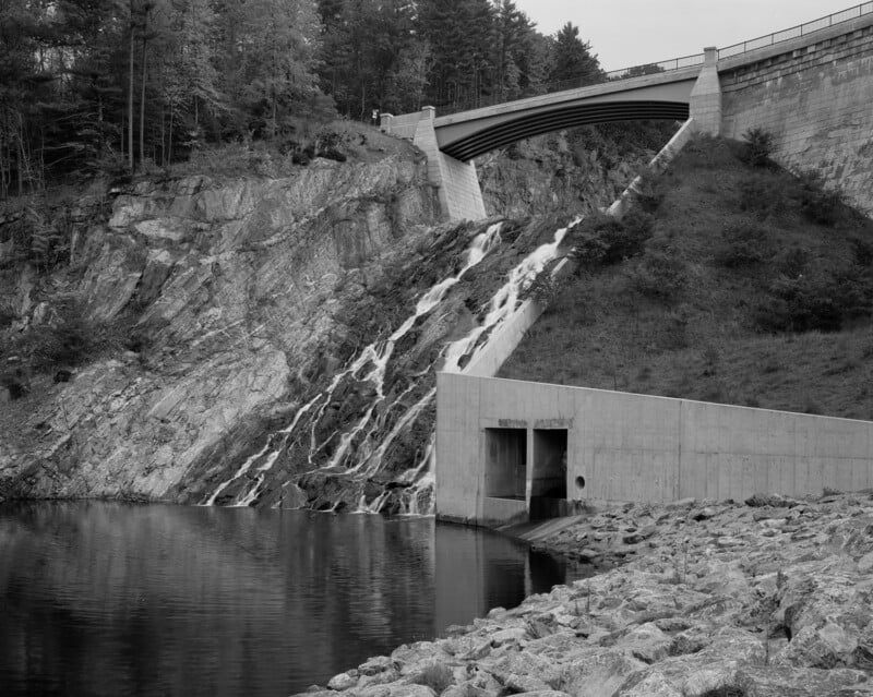 A concrete dam bridge spans a rocky hillside, with water cascading down spillways into a calm reservoir below. Trees line the top of the hill, and rocks cover the shoreline in the foreground. The image is in black and white.
