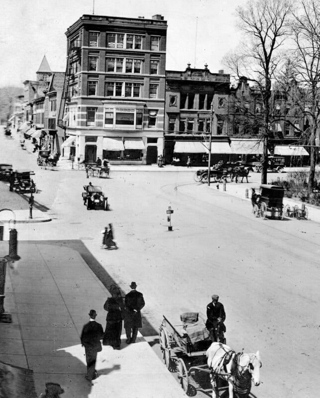 Historic black-and-white photo of a city street with horse-drawn carriages, early automobiles, and people walking on sidewalks near multi-story brick buildings and storefronts. Leafless trees line the street.