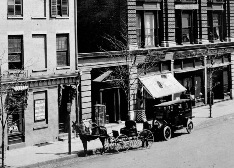 A black-and-white photo of an early 20th-century city street shows a horse-drawn carriage and an old-fashioned car parked in front of storefronts with awnings and signs. Leafless trees line the sidewalk.
