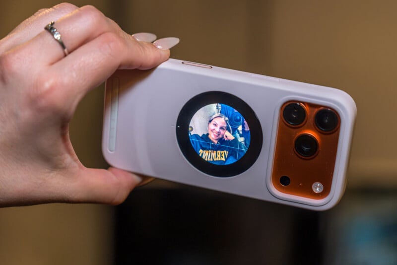 A hand holding a smartphone with a circular display on the back, showing a smiling person taking a selfie. The phone has a white case and three camera lenses visible.