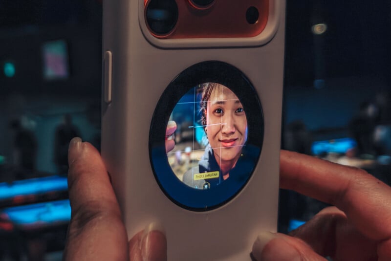 Close-up of a smartphone held in both hands, showing a smiling woman's face on the lens or circular screen in the center of the phone, with a crosshair overlay and a blurred background.