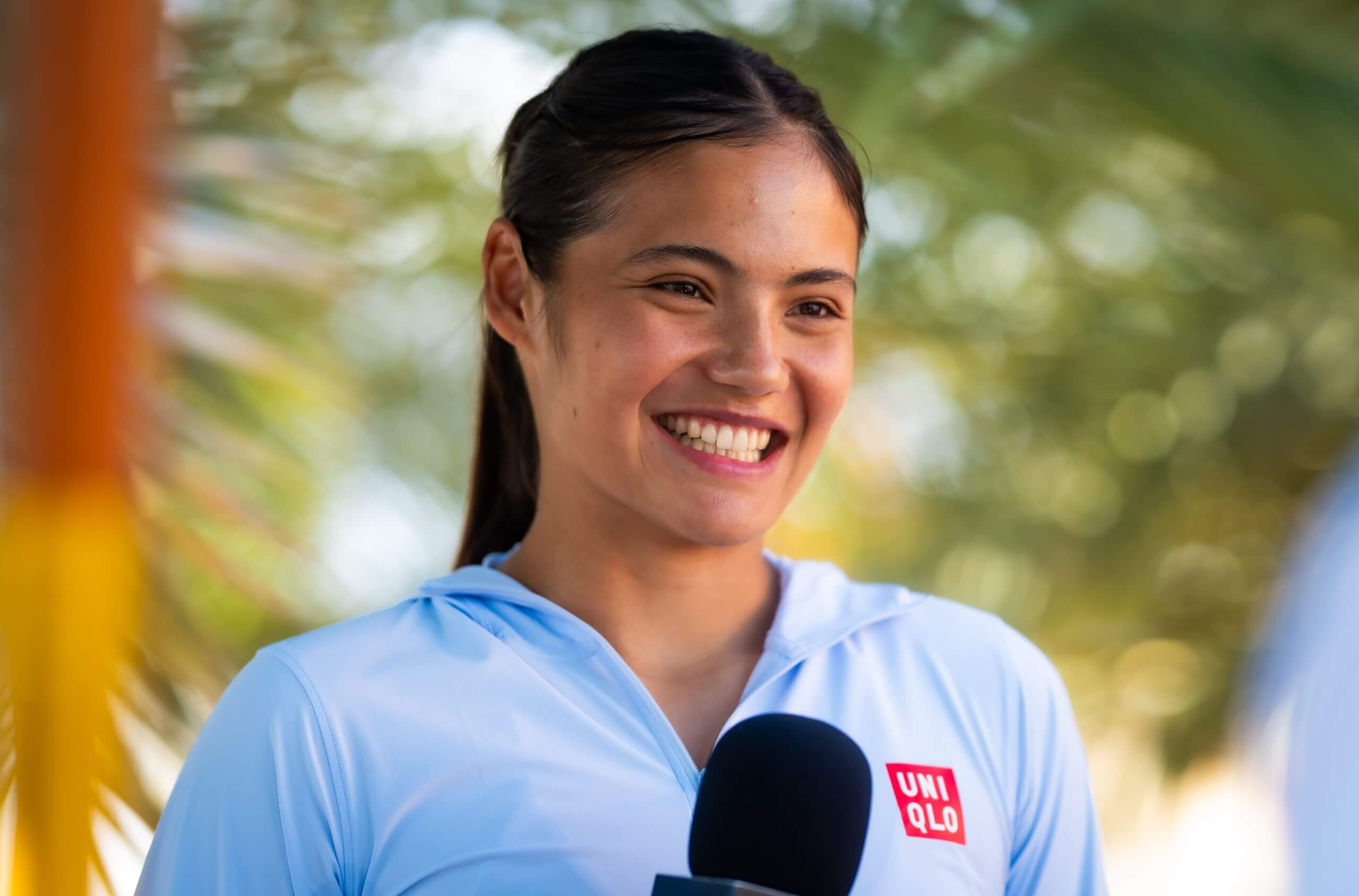 Emma Raducanu smiles while wearing a light-blue tennis outfit with a Uniqlo logo.