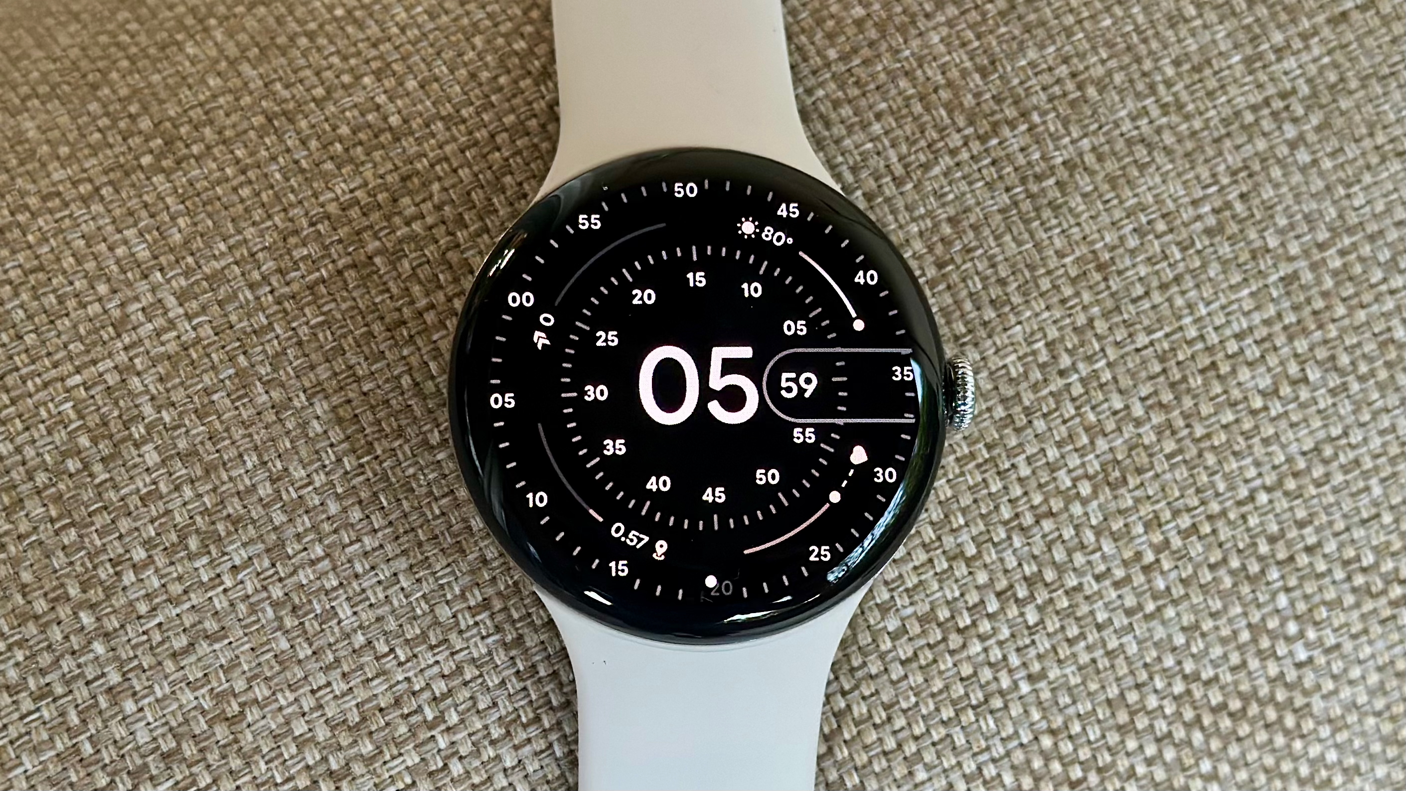 A photo of the Google Pixel Watch 4 sitting on a cloth chair cushion, showing the Concentric watch face with white hour, minute, and second numbers extending to the edge of the domed display.