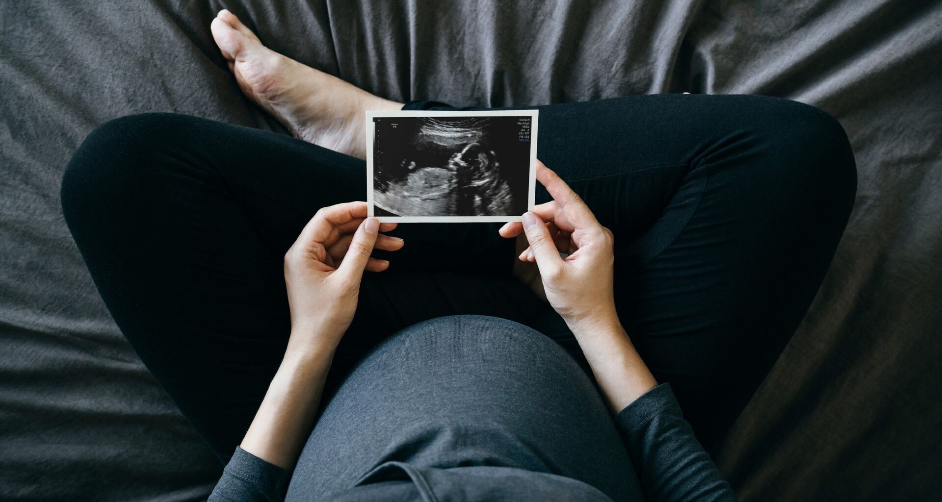 A top down shot of a pregnant person wearing a blue shirt and black pants sitting cross-legged on a blue bed, holding an ultrasound image in front of them.