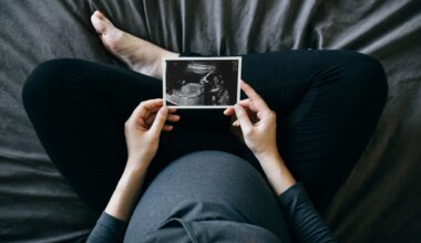 A top down shot of a pregnant person wearing a blue shirt and black pants sitting cross-legged on a blue bed, holding an ultrasound image in front of them.