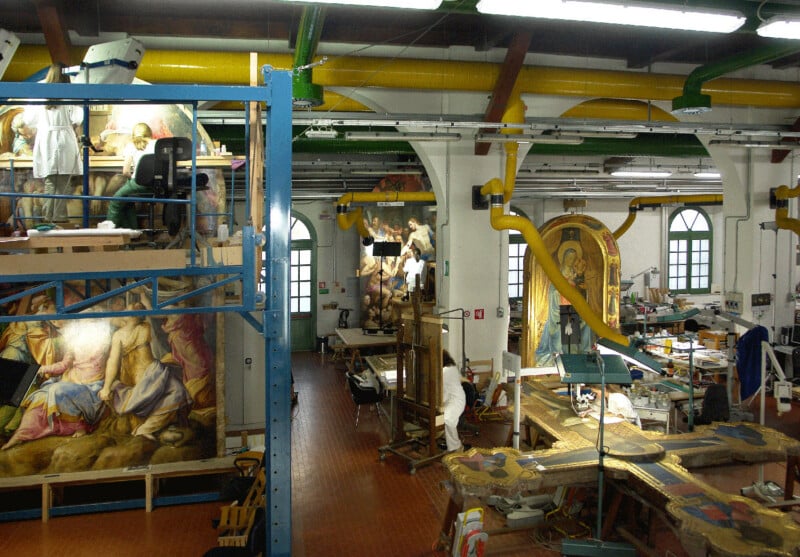 Large art restoration studio with high ceilings, filled with conservation equipment, large religious paintings, and artifacts being restored. Two people work on scaffolding and tables amid the artworks and restoration tools.
