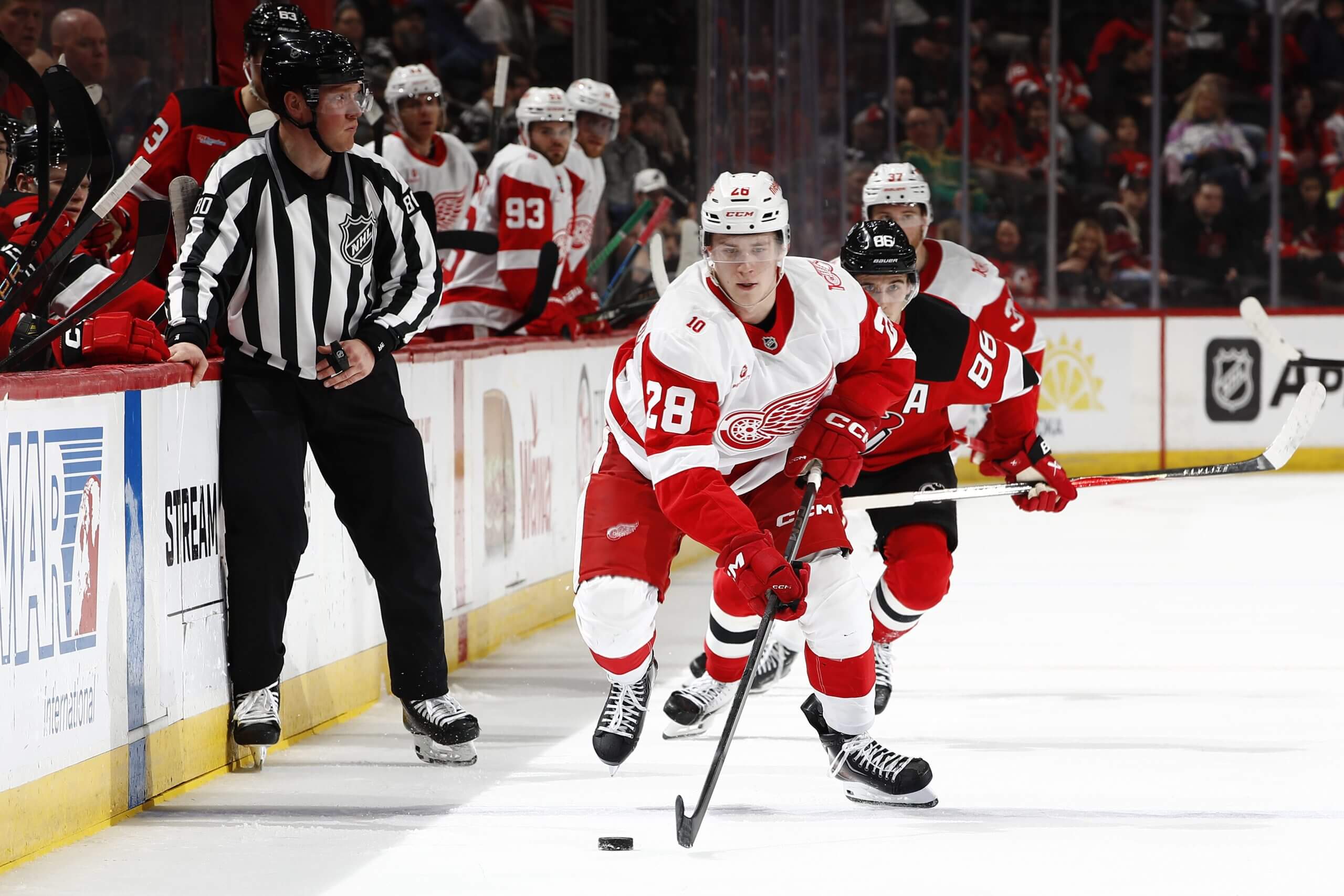 Michael Brandsegg-Nygaard skates past a referee with the puck during a Red Wings game, with a Devils player barely visible over his shoulder.