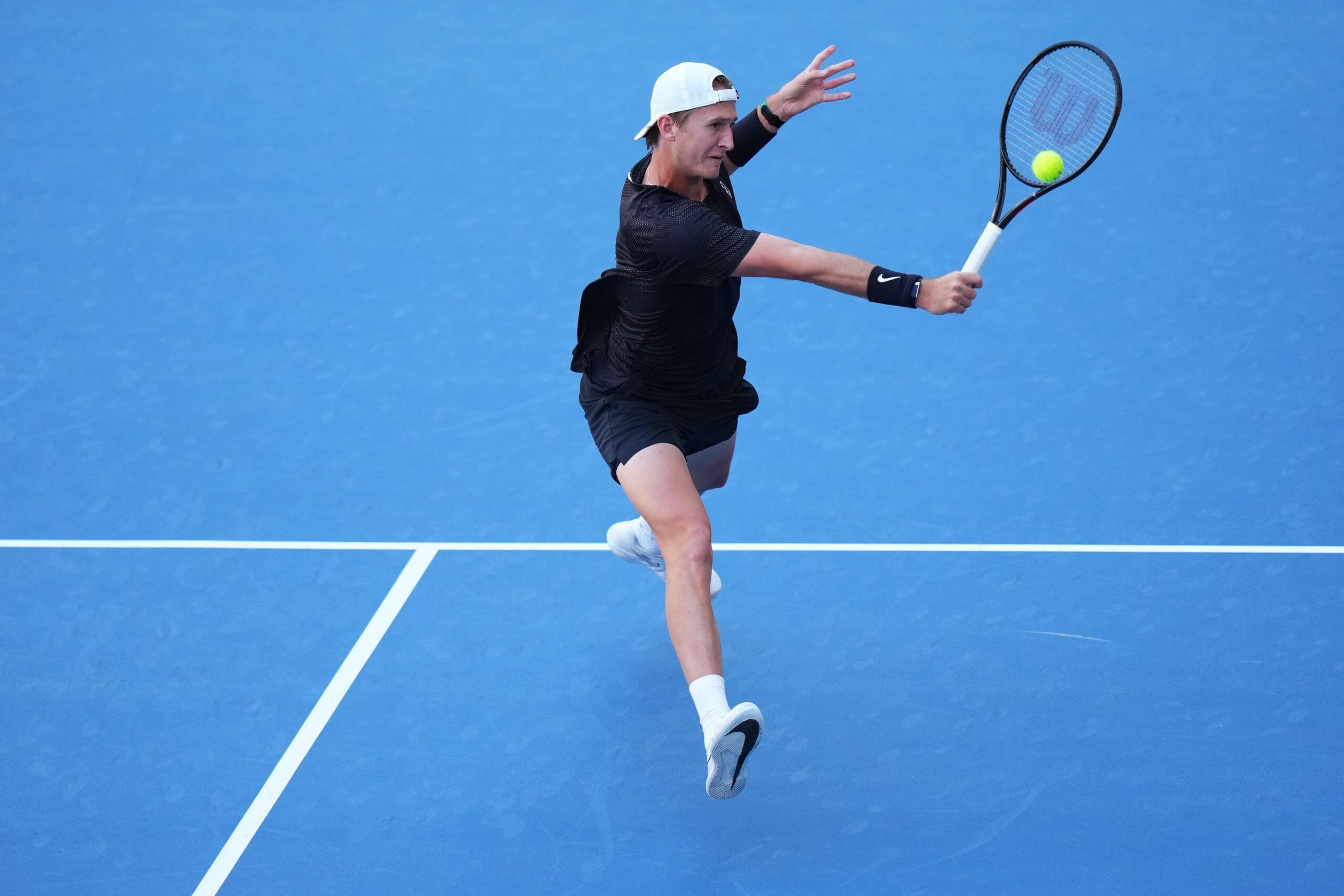 Sebastian Korda, in a dark top and wearing a white ballcap backward, makes a backhand shot against Carlos Alcaraz during their match at the Miami Open at Hard Rock Stadium.