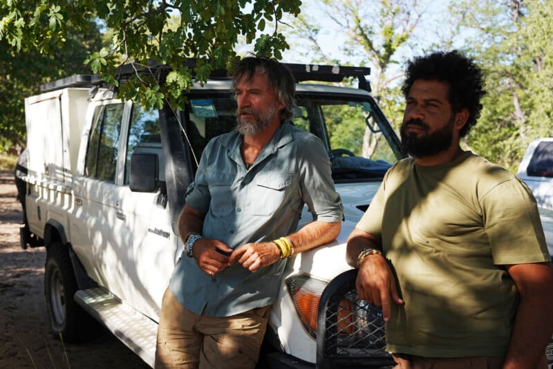 Two men stand beside a white off-road vehicle under a tree, looking thoughtfully into the distance. One wears a light blue shirt and the other a green t-shirt. Sunlight filters through the leaves above.