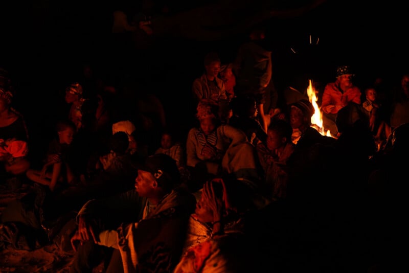 A group of people sit closely together at night, illuminated by the warm glow of a campfire. Their expressions appear somber and reflective, and the surrounding area is shrouded in darkness.