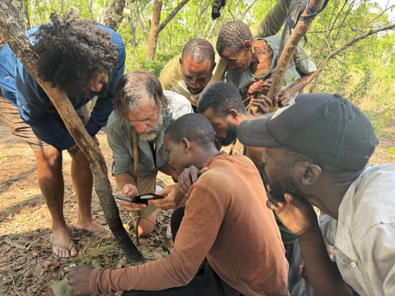 A group of seven men gather closely in a wooded outdoor setting, intently looking at a smartphone held by one of them. Sunlight filters through the trees, and the ground is covered in dry leaves and branches.