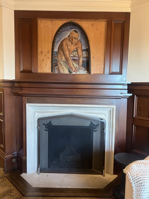 A wooden-paneled fireplace with a marble surround, featuring a painting of a person working with dough above the mantel.