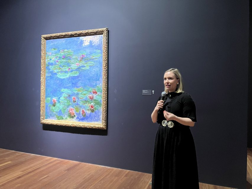 A woman with a microphone stands next to a framed painting of water lilies displayed on a dark wall in an art gallery.