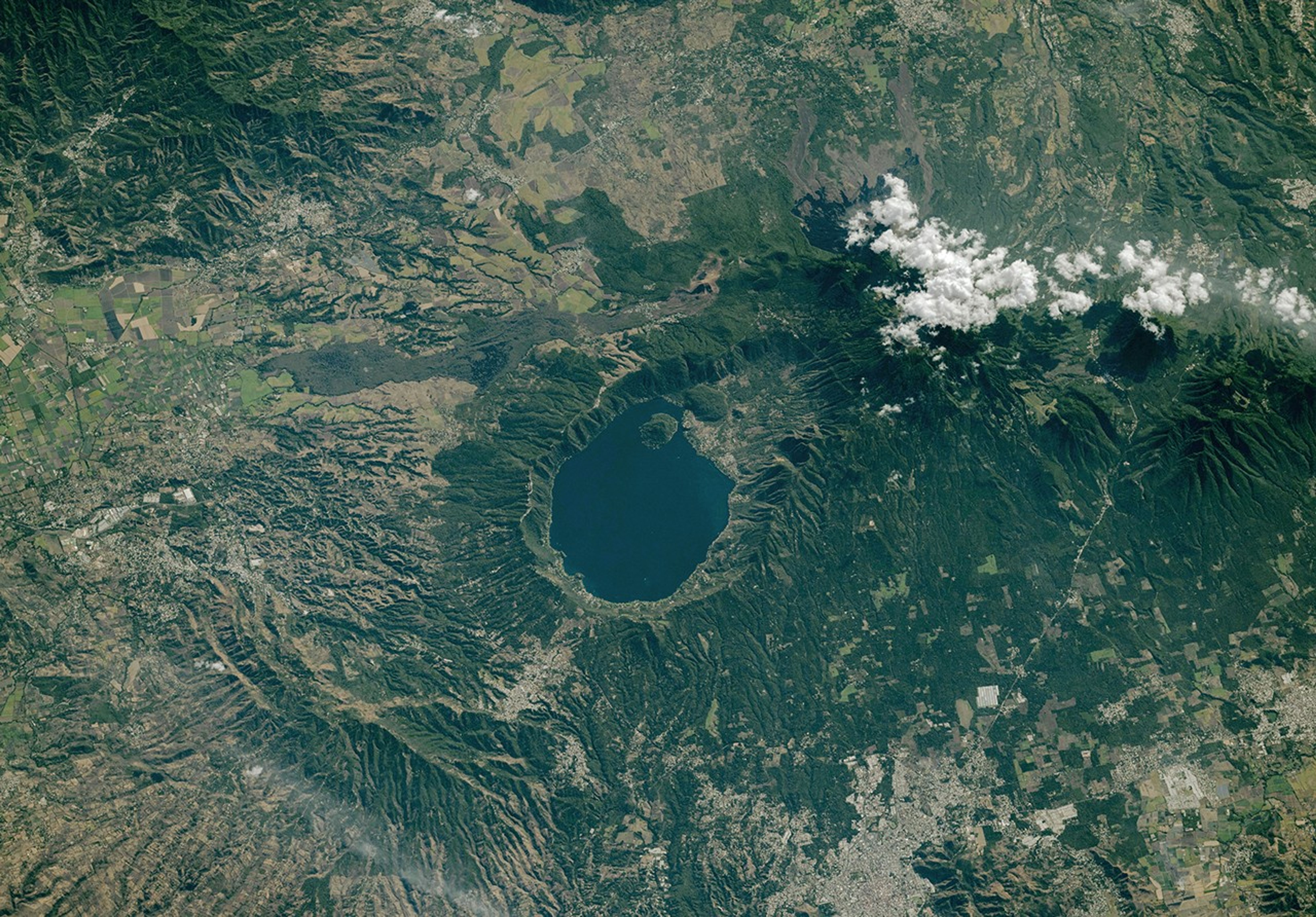 A blue lake rests within a caldera with steep walls. Several volcanoes near the caldera are capped by clouds. The terrain is mostly lush and green, with patches of gray urban areas.
