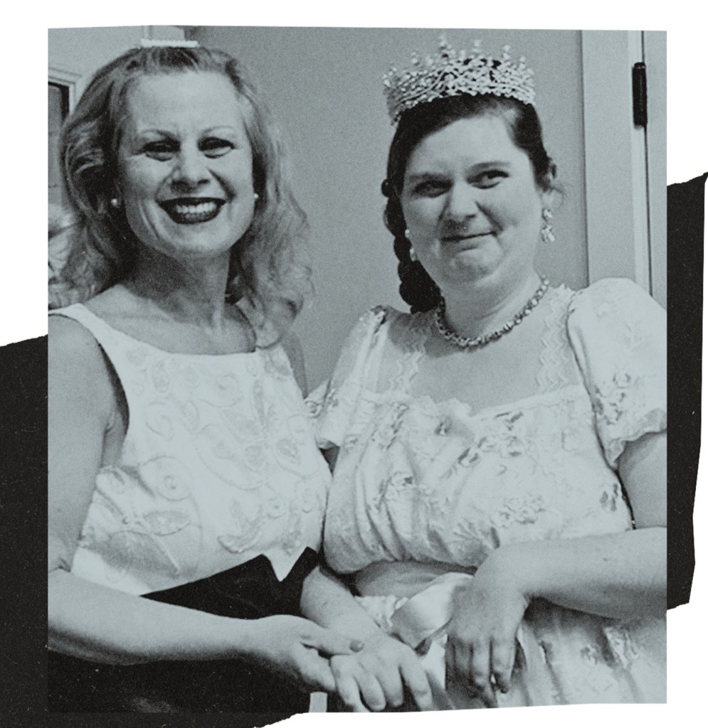 Black and white photo of two women standing side by side and smiling. One wears a sleeveless dress, the other wears a light-colored formal gown, a tiara and a necklace. They are holding hands. The image is set against a light background with a dark textured shape behind it.