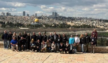 A group of people pose for a photo on a stone platform overlooking Jerusalem, with the Dome of the Rock and cityscape visible in the background under a partly cloudy sky.