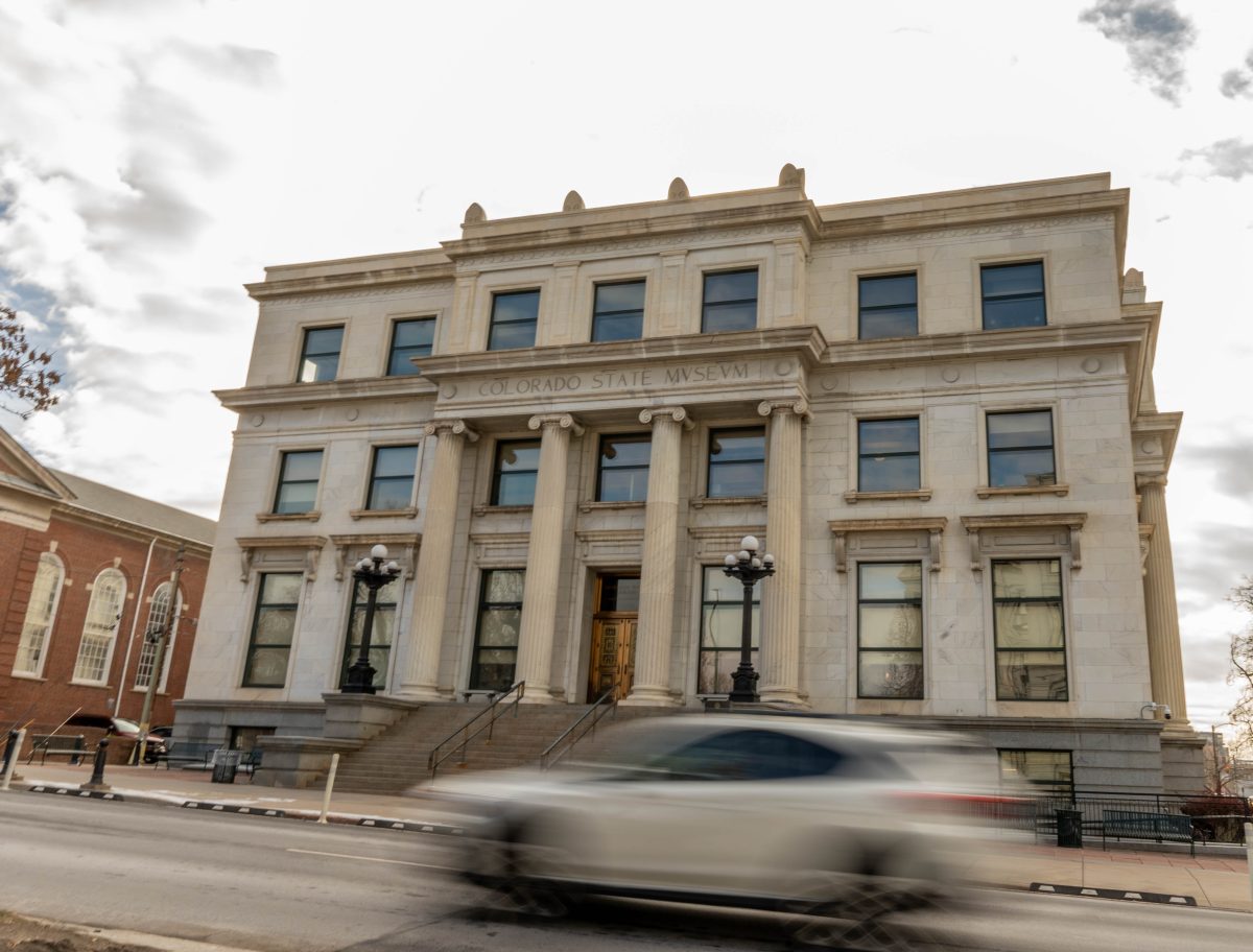 A three-story classical building labeled "Colorado State Museum" with tall columns and large windows. A white car drives by on the street in front.