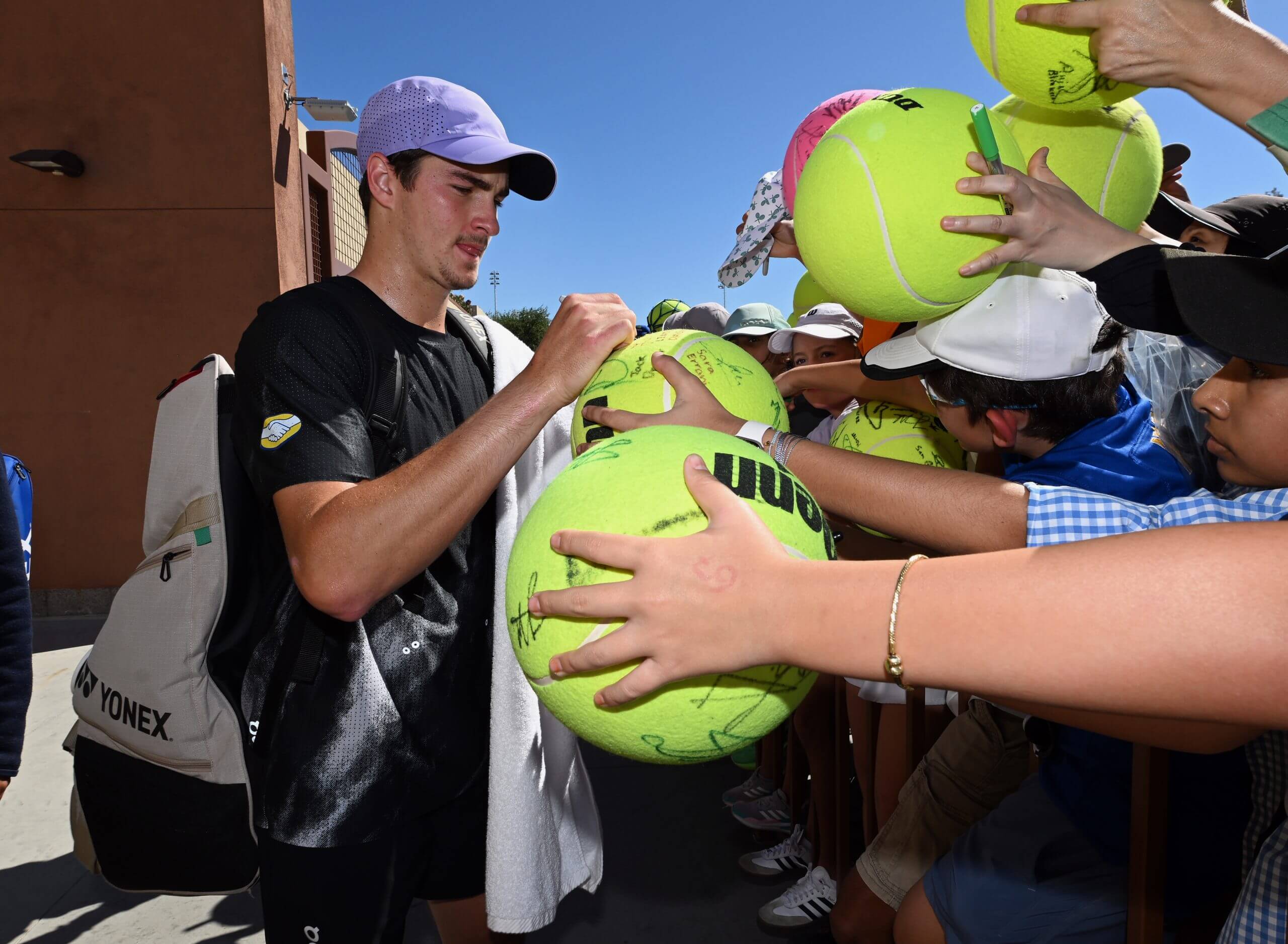 João Fonseca signs jumbo tennis balls in outstretched hands, in front of a brown building under a blue sky.