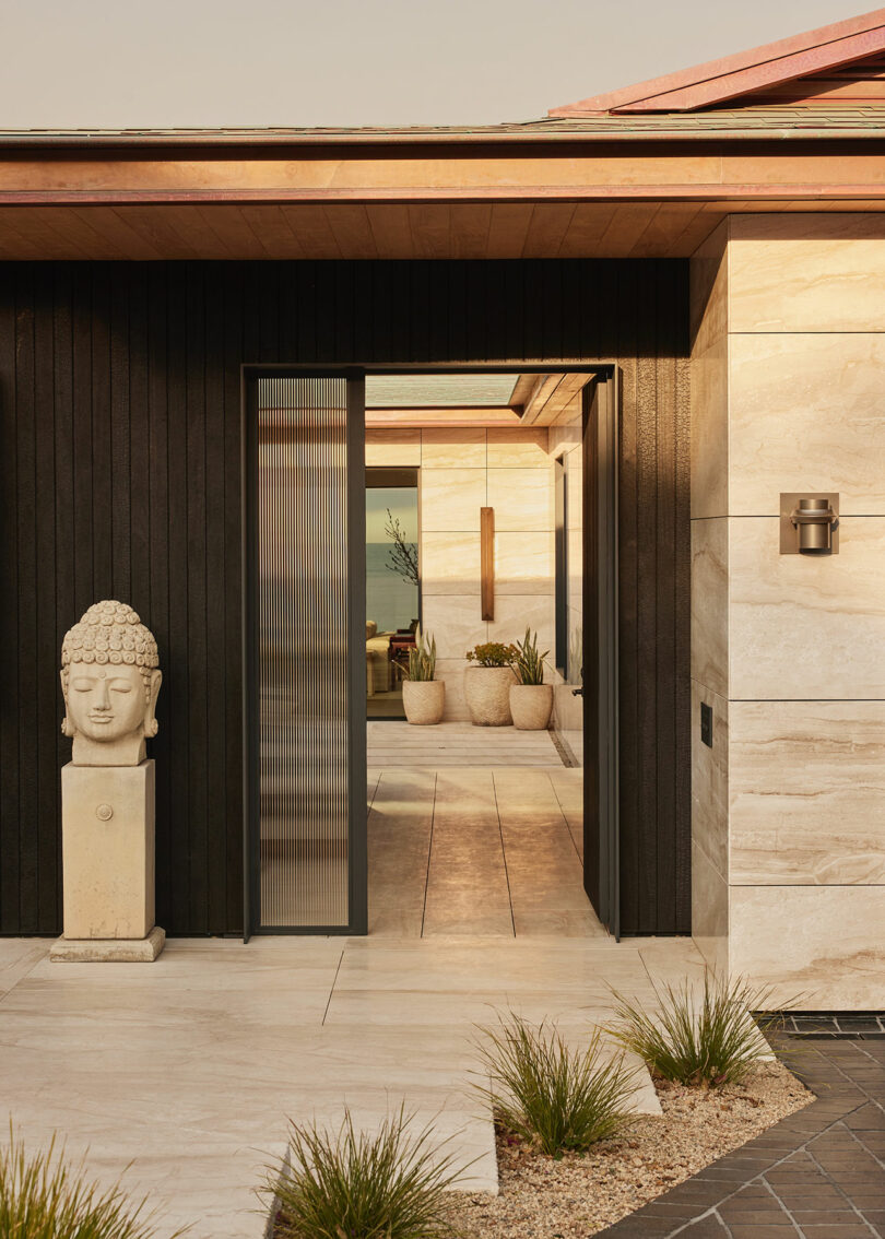Modern house entrance with light stone and dark wood paneling, a Buddha head statue on a pedestal, potted plants, and a partially open glass door.