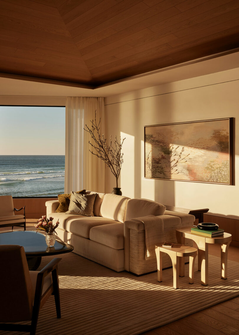 A sunlit living room with a beige sofa, a large seaside window, minimalist furniture, a wall painting, and a vase with branches. Ocean view in the background.
