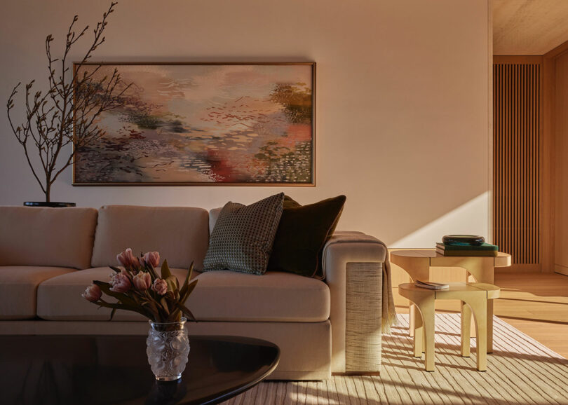 A beige sofa with green cushions sits below a large abstract painting; a side table, books, and vase with pink flowers complete the warmly lit living room scene.