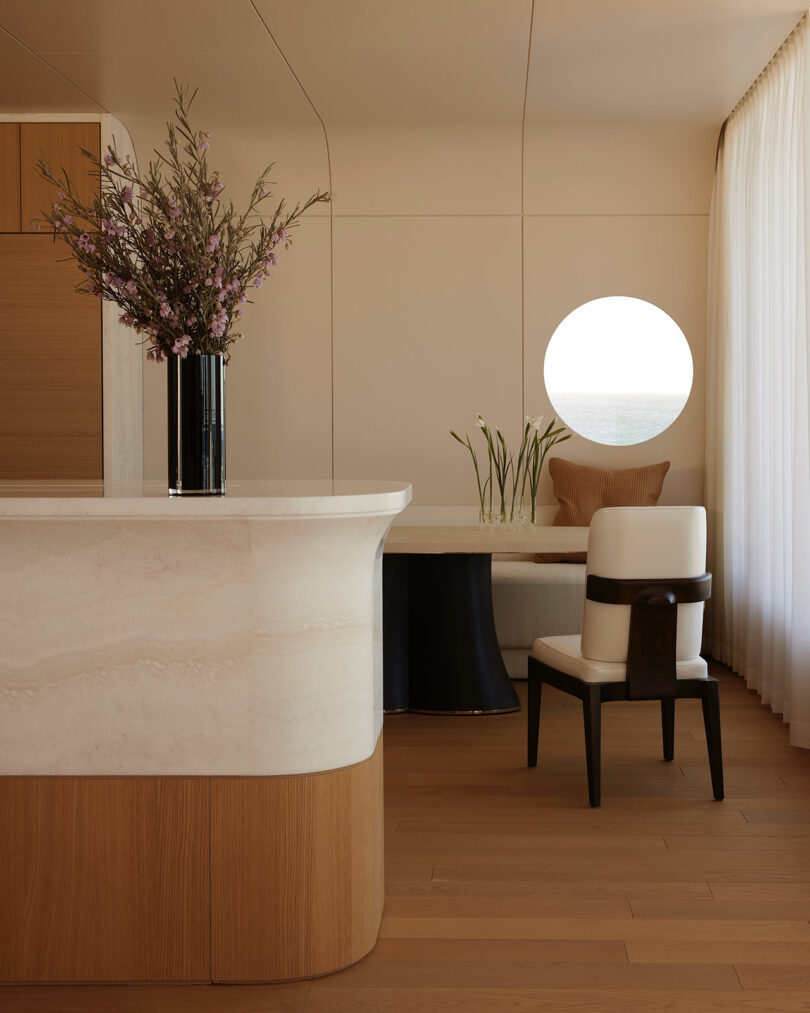 Minimalist dining area with a curved stone counter, black vase of flowers, round table, white chair, and a circular window with sheer curtains and a beige cushion.