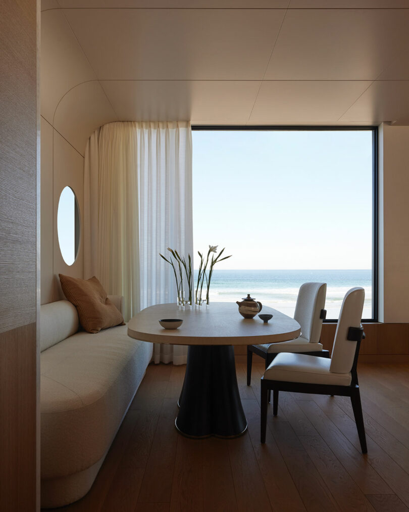 Minimalist dining area with a round table, two chairs, a sofa, and a large window offering a view of the sea; neutral tones and natural light fill the room.