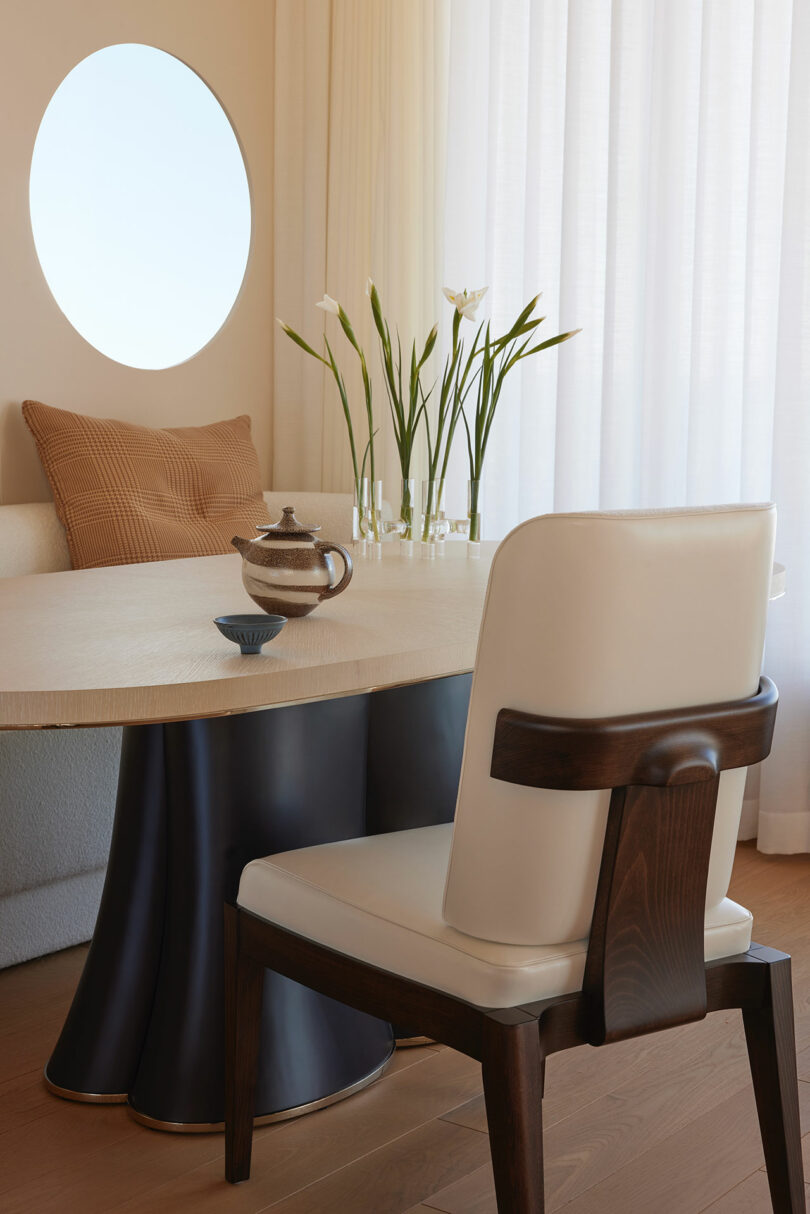 A modern dining area with a round table, a white cushioned chair with dark wood accents, a beige sofa, a round wall mirror, and vases with flowers on the table.