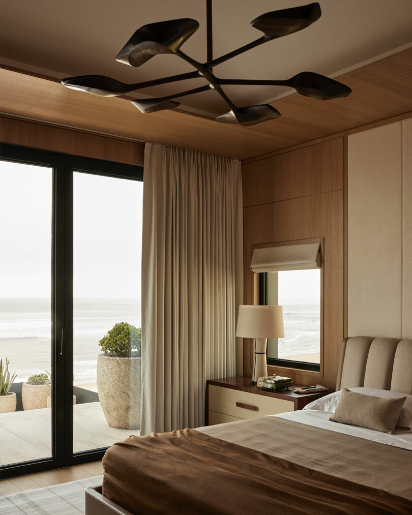 Modern bedroom with a large bed, wooden walls, a stylish black ceiling light, bedside lamp, and sliding glass doors opening to a balcony with an ocean view.
