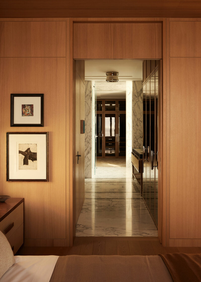 A wooden-paneled bedroom doorway opens to a marble hallway with framed art on the wall and natural light illuminating the corridor beyond.