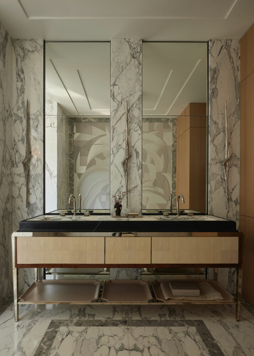 Modern bathroom with a marble countertop, dual sinks, large mirrors, wall-mounted lighting, and marble flooring; shelves below the vanity hold towels and decor.