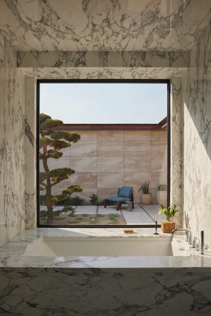 A marble-walled bathroom with a bathtub faces a large window overlooking a minimalist patio with a manicured tree, plants, and a blue outdoor chair.