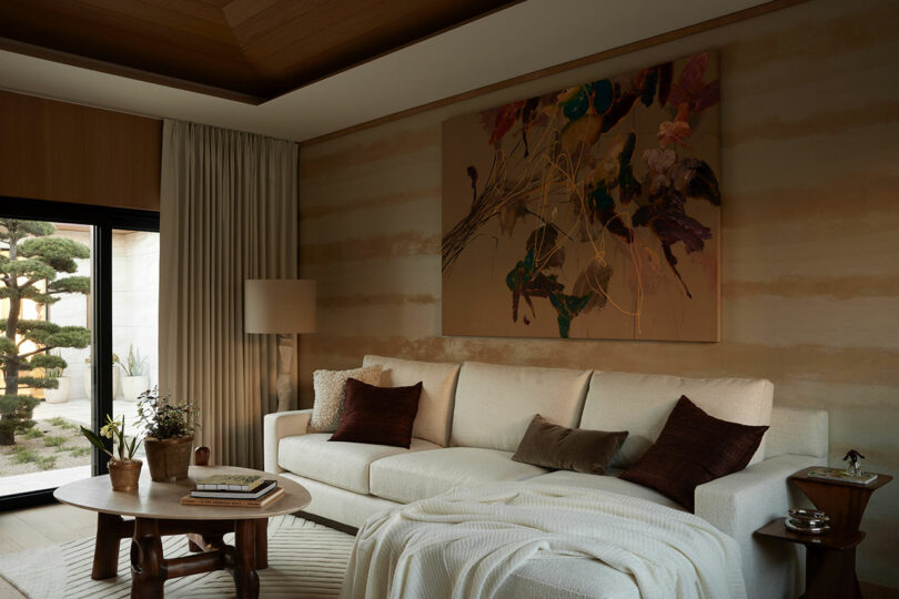 A modern living room with a cream sofa, throw blanket, brown cushions, wooden coffee table, abstract wall art, lamp, and large window with garden view.