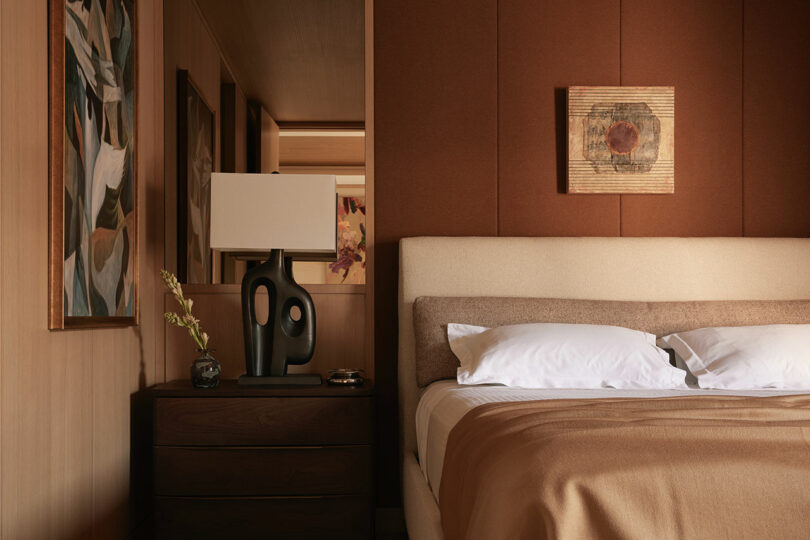 A neatly made bed with beige and brown bedding sits beside a dark wood nightstand with a modern black lamp, glass dish, and a vase with a single stem; abstract art hangs on the walls.