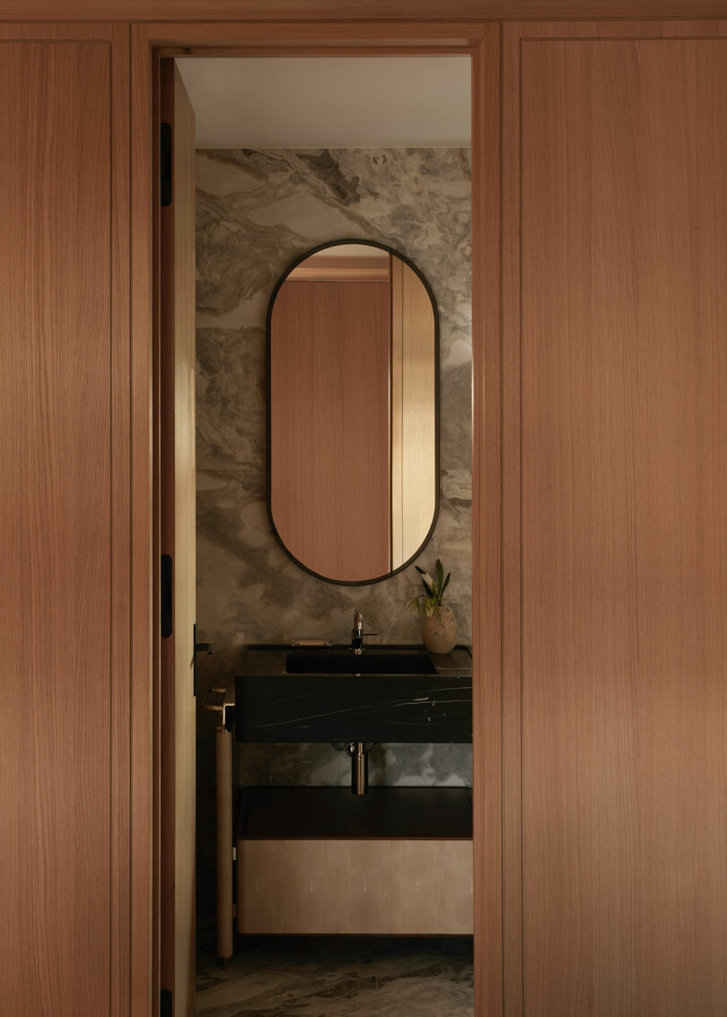 A modern bathroom with light wood walls, a black countertop and sink, an oval mirror, and a small potted plant, set against a marbled stone wall.