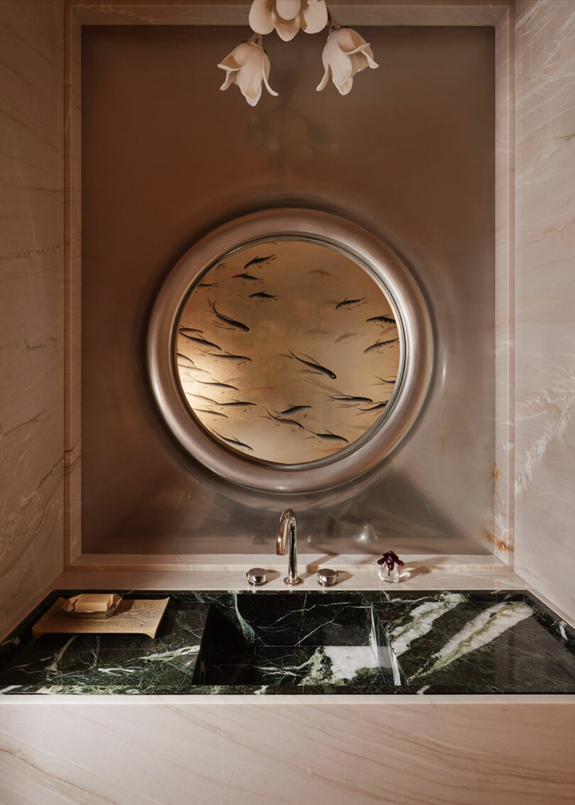 A modern bathroom sink with a dark green marble countertop, metal faucet, and a round mirror featuring a fish pattern, set against light-colored marble walls.