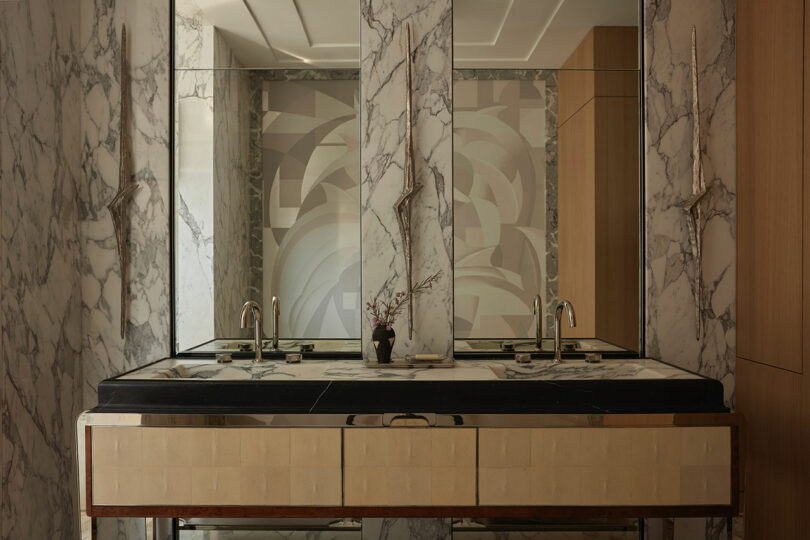 Modern bathroom with marble walls, double sinks, a large mirror, and wood accents; a small plant in a vase sits between the sinks.