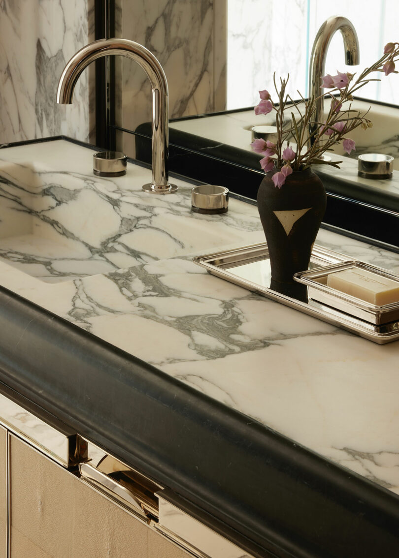 A marble bathroom countertop with a modern chrome faucet, a black vase with pink flowers, and a soap dish beside a large mirror.