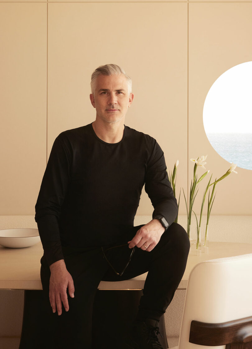 A man with short gray hair wearing a black long-sleeve shirt sits on the edge of a light-colored table next to a vase with white flowers, in a minimal modern room with a round window.