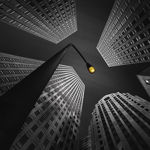 A black and white photo of tall skyscrapers viewed from the ground, converging toward the sky, with a single street lamp in the center emitting a yellow light.