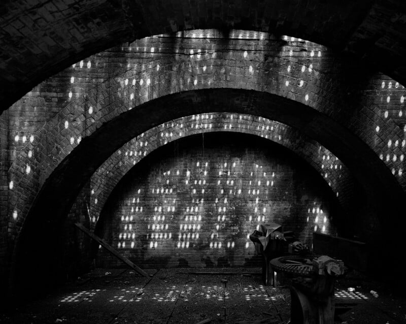 Sunlight streams through small holes in a brick arched ceiling, casting a pattern of bright dots onto the walls and floor of a dark, empty, and dusty underground room with scattered debris.
