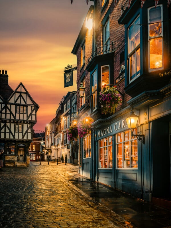 A picturesque cobblestone street at sunset, lined with historic buildings and glowing shopfronts. Hanging lanterns and potted flowers decorate the scene, creating a warm, inviting atmosphere.