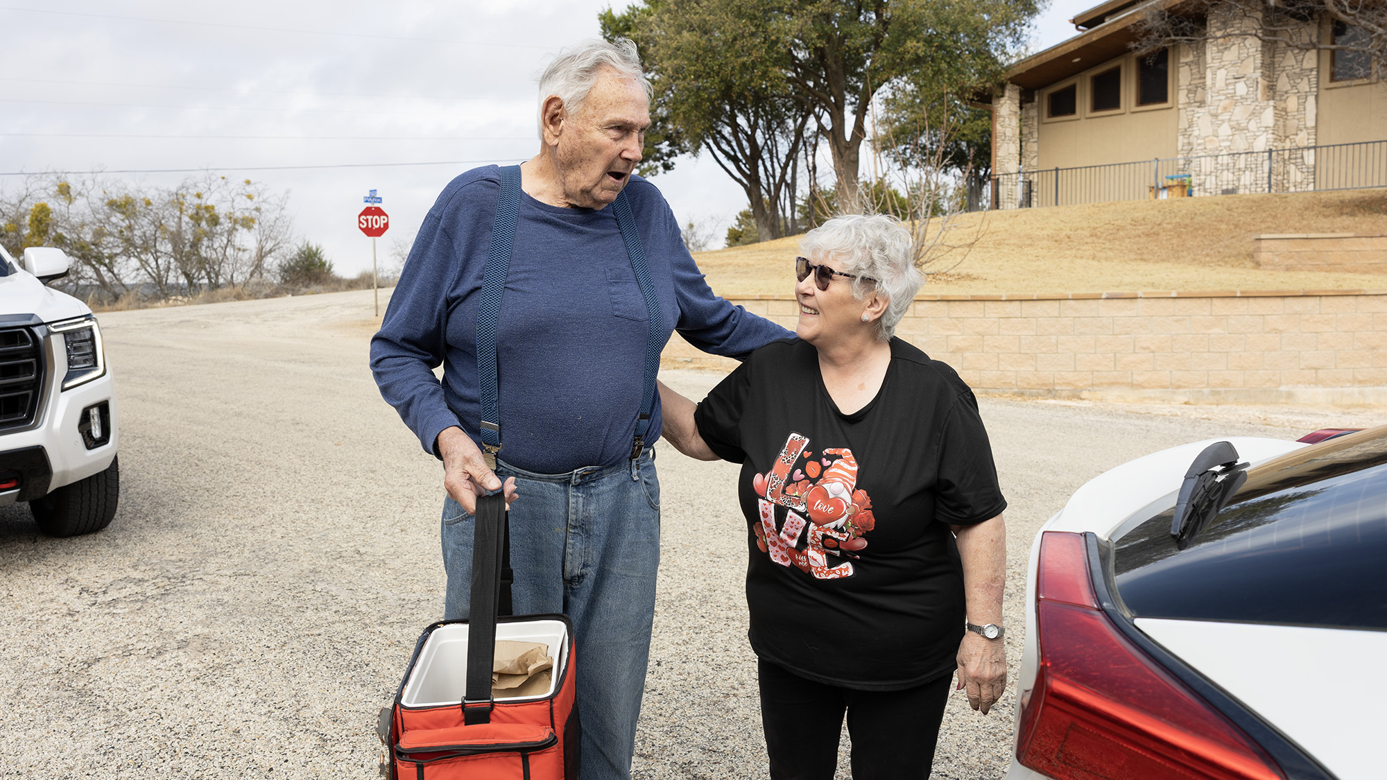 An older man and woman stand with their arms on each other's shoulders, next to a parked car. The man holds a cooler full of packed meals.
