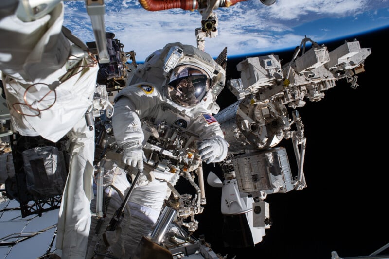 An astronaut in a spacesuit works outside the International Space Station with Earth and space visible in the background. Various equipment and station parts surround the astronaut.