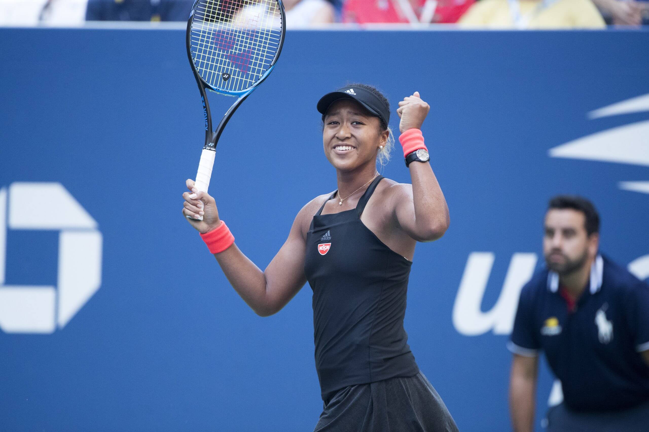 Naomi Osaka raises her racket and pumps her fist, wearing a black tennis outfit.