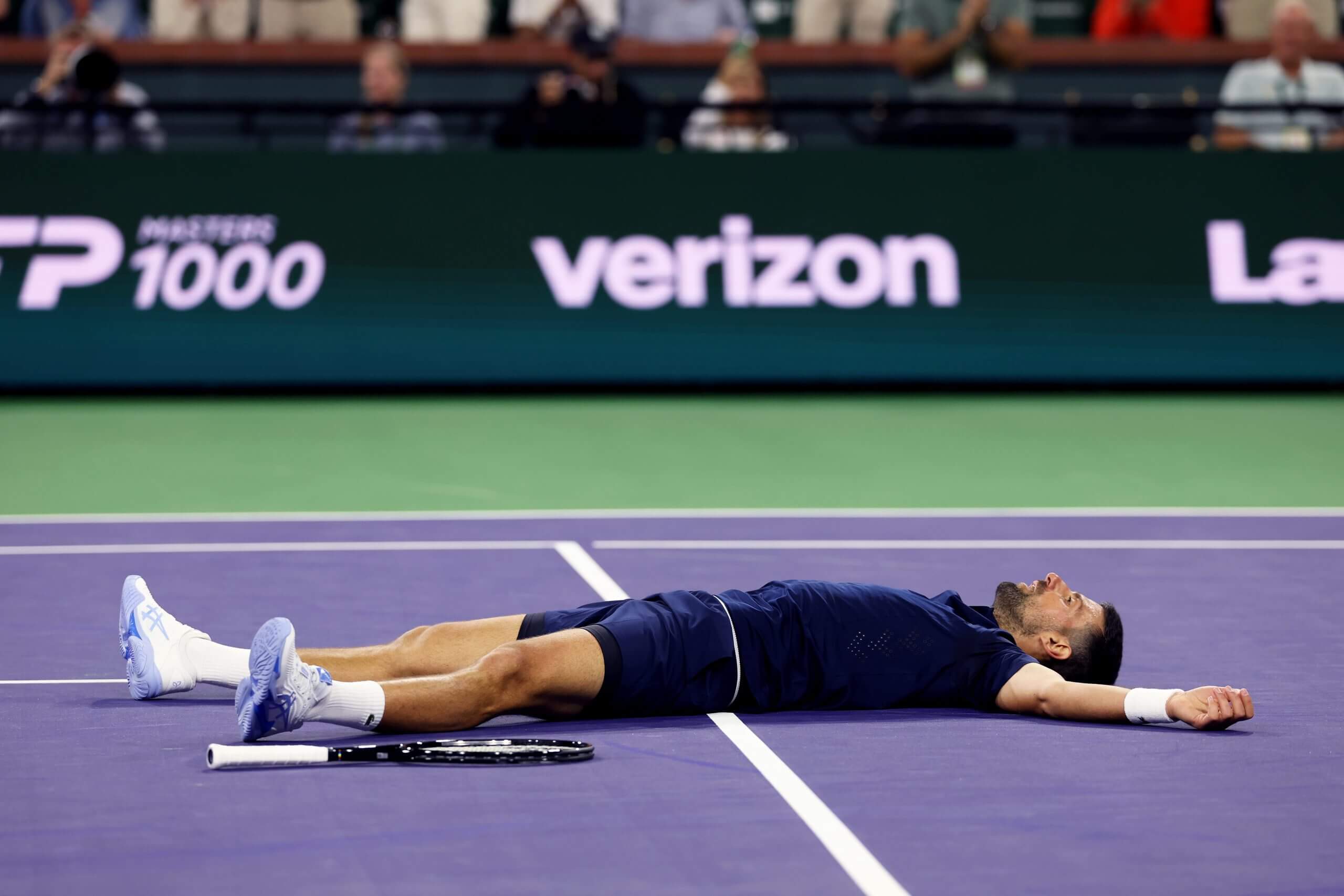 Novak Djokovic lies flat on his back on a purple tennis court, his tennis racket at his side.