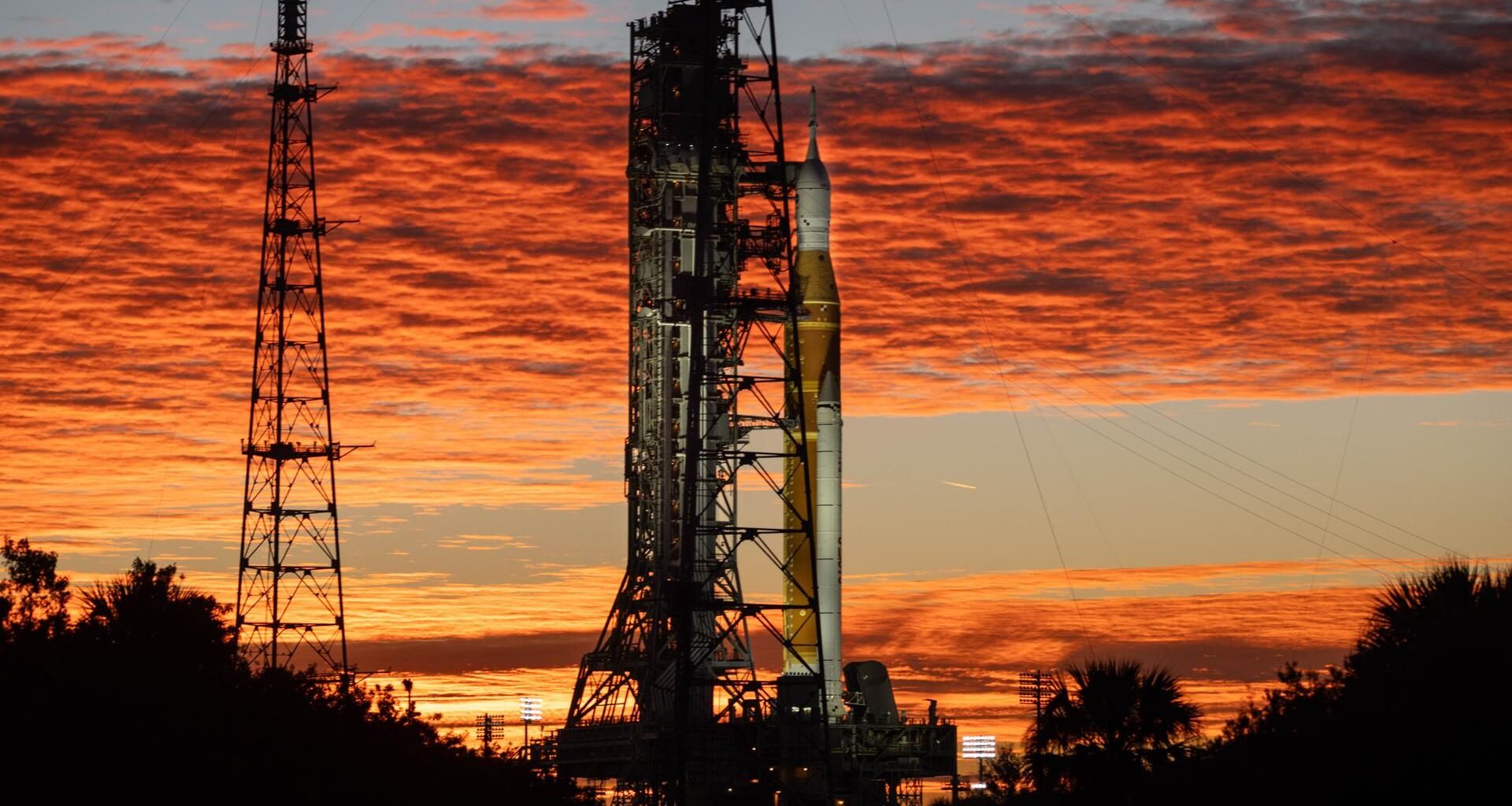 An orange rocket with white side booster stands next to the launch tower during a colorful sunrise.
