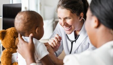 Woman doctor with dark-haired ponytail, white lab coat and stethoscope examines baby with mother and teddy bear on either side of baby