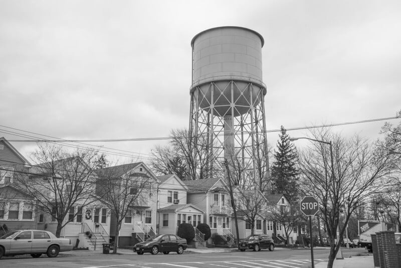 A large water tower rises above a residential neighborhood with several houses, parked cars, bare trees, and a stop sign at a street intersection, all under an overcast sky.