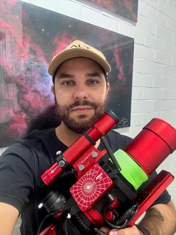 A man wearing a beige cap holds a red telescope and smiles at the camera. Behind him, there is a colorful photograph of a nebula and a white brick wall.